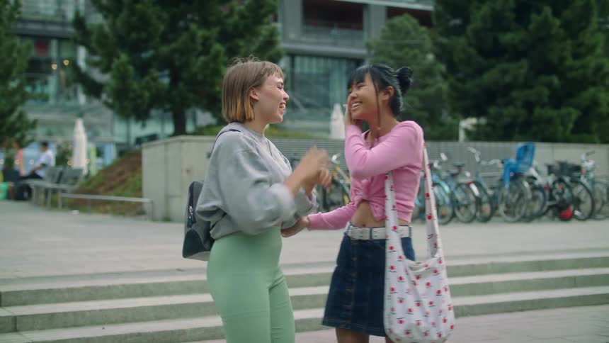 A young woman sneaks up behind another woman and surprises her with a kiss before they embrace