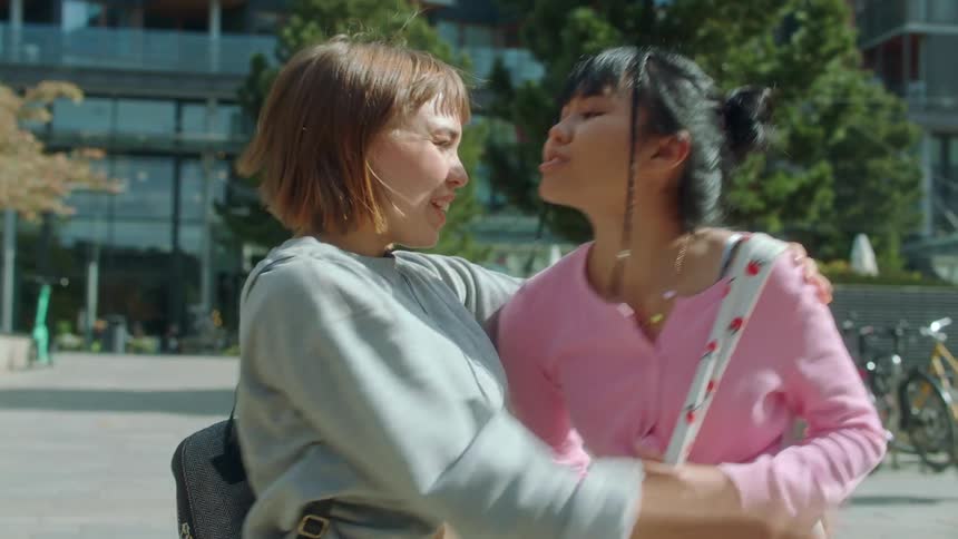 A young woman walks up behind another one and gives her a kiss and hug