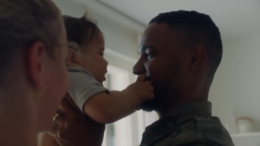 Fatherin military jacket holds child in air then kisses him before turning to look at his wife