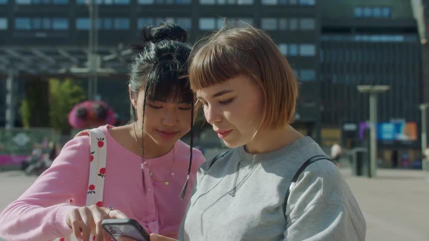 Two girls are together on a downtown street and one girl is showing the other something on her phone