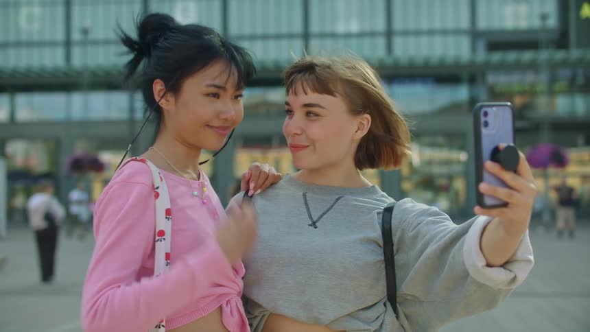 Friends smile with their arms around each other and take selfie as pedestrians approach store entrances