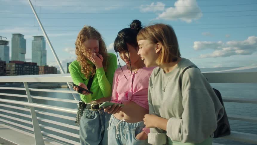 Three girl friends lean against a bridge railing laughing and looking at their phones
