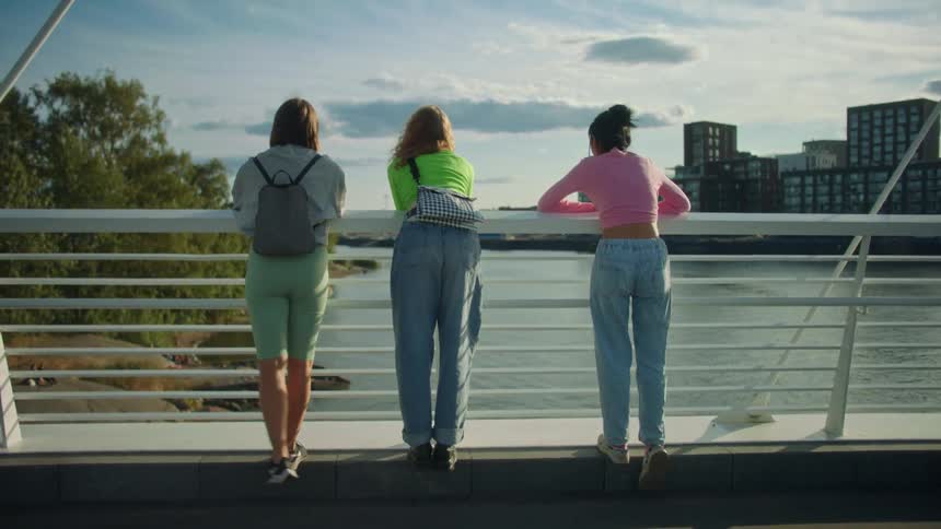 Three women standing on a bridge with railing with a river buildings the sky and clouds in the background