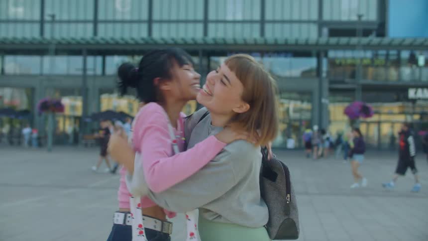 Female friends hug and stand talking in plaza as pedestrians move around entrance of building with large screen on the side