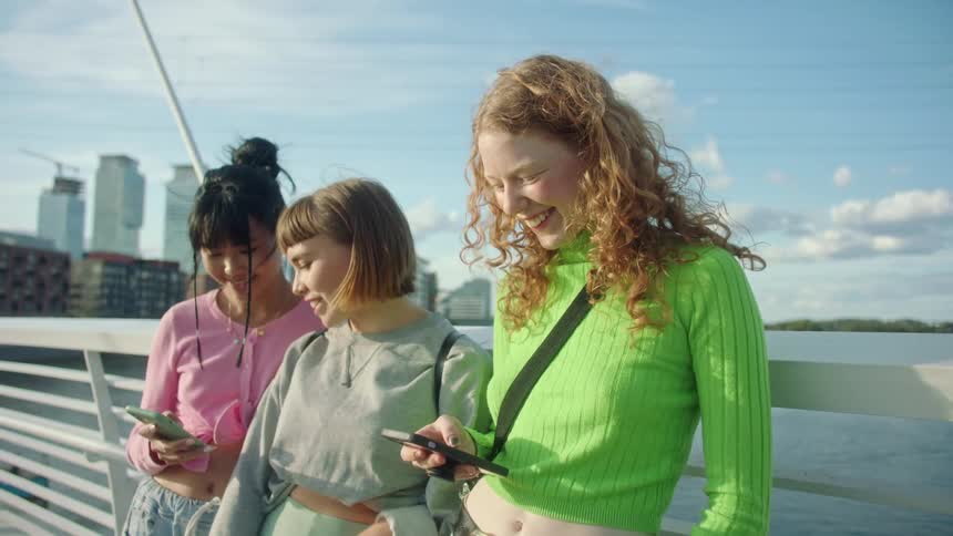 A trio of young women stand together while looking at their smartphones while smiling and laughing