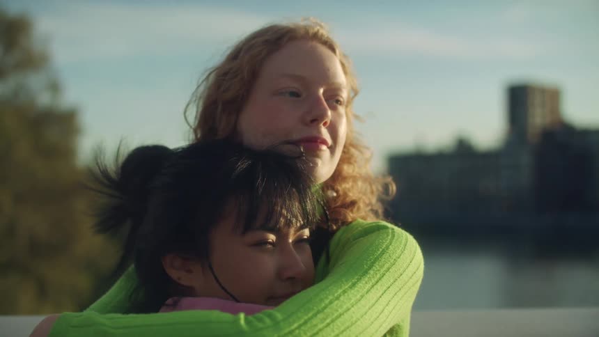 A woman hugs her girlfriend and pulls her in close as they stand on a bridge near downtown