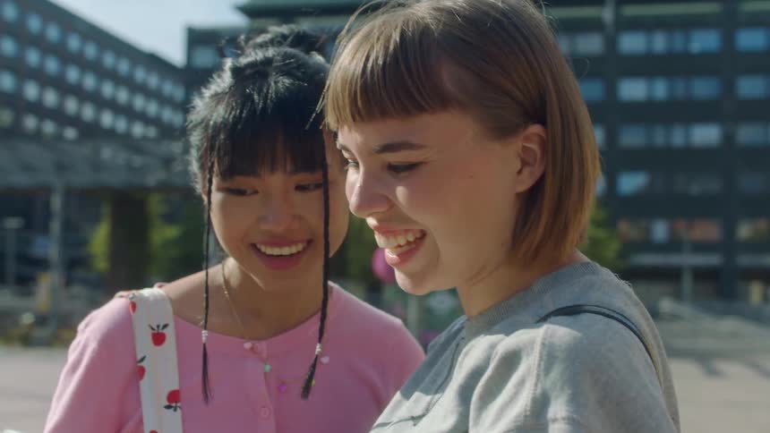 Two young women chat with someone via video call on their phone while standing downtown