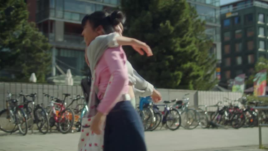 Female friends laugh and hug and dance holding hands standing near parked bicycles while a pedestrian and a man on a scooter pass