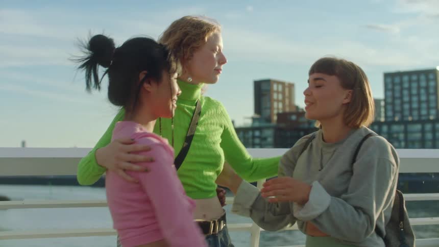 Three girls are talking and give each other a group hug with a city and lake in the distance