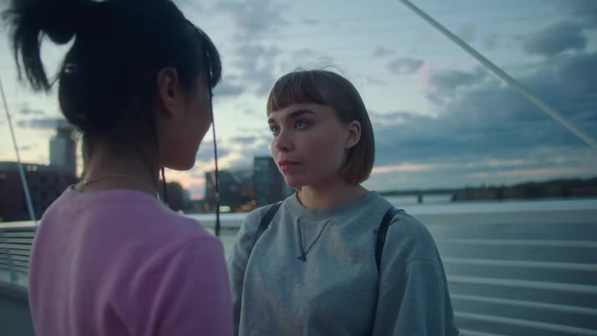 A pair of young women embrace while standing on a bridge in the evening