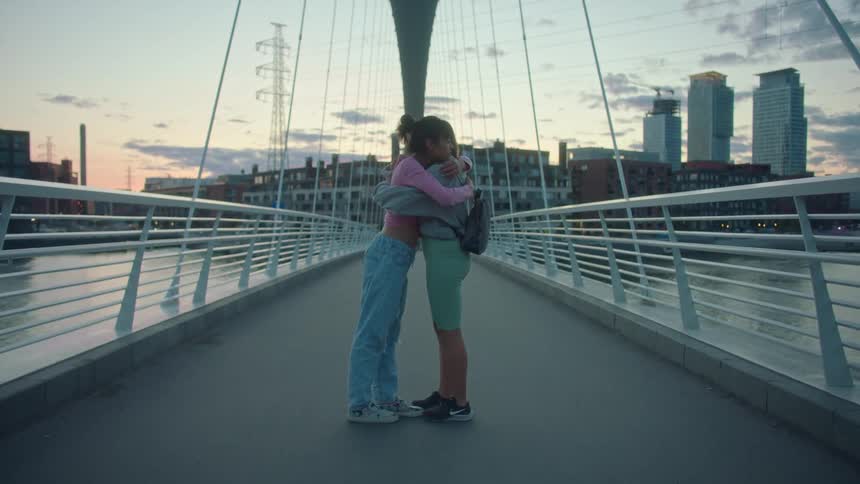 Two girls standing on a pedestrian bridge lean forward and hug each other
