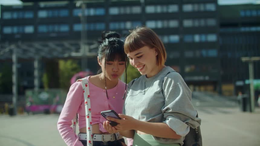 Two women are standing outside in front of a building and looking at a phone