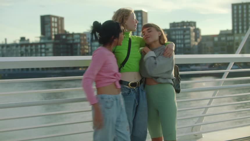 A group of young women on a bridge over a river hug one another before waving farewell as one woman walks away