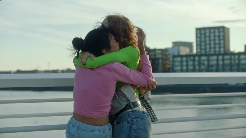 Three woman on a bridge hugging with buildings the sky and clouds in the background