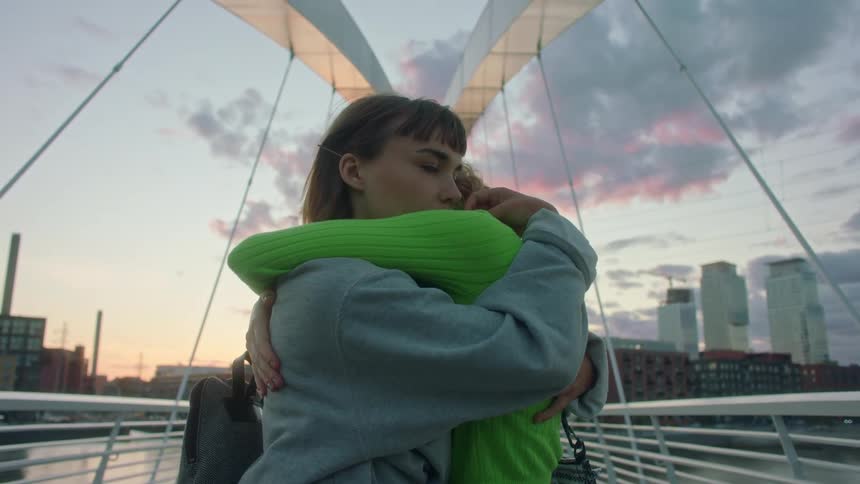 Two teenage girls hugging on a city bridge at sunset
