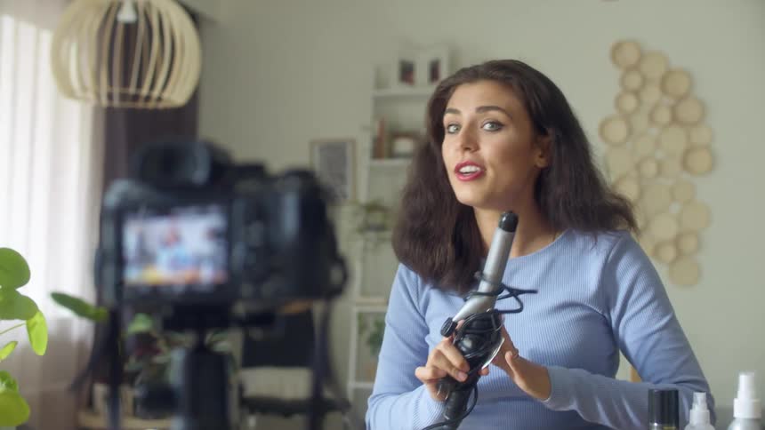 A woman in a room brushing her hair with a brush while talking to a camera with a wall and window in the background