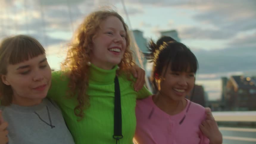 Three women walking and hugging with buildings the sky and clouds in the background