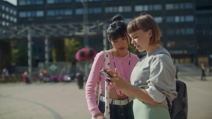 A young woman uses her smartphone as another young woman watches while they stand together outdoors