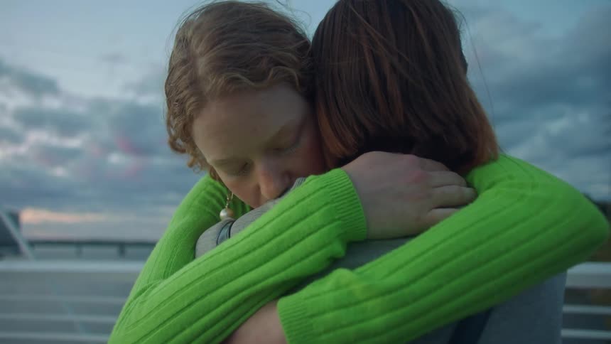Two teenaged girls hug with a lake and city buildings in the distance under a cloudy sky