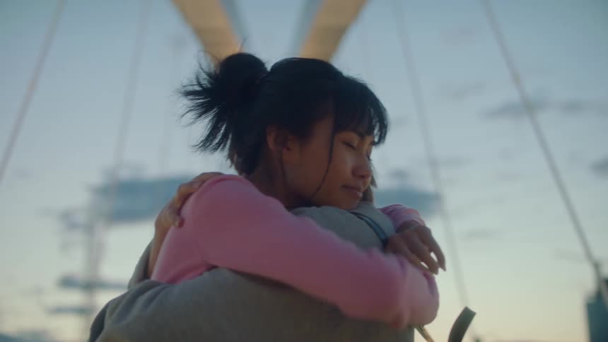 Two women on a bridge hugging with buildings the sky and clouds in the background