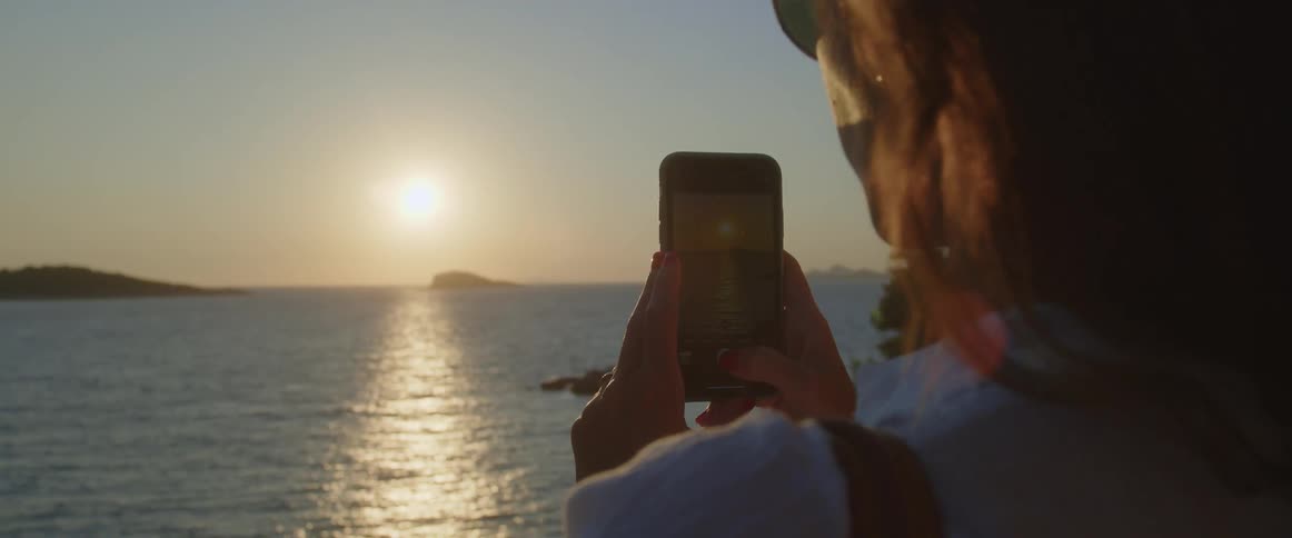 A woman takes a photo with her phone of the sun setting over the sea