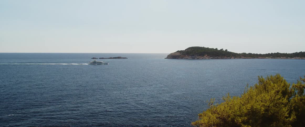 Ship cruises into cove nearing tree covered promontory under light blue sky
