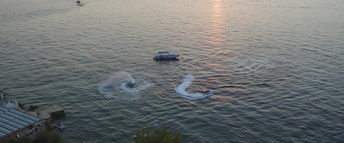 People ride jet skis over water between dock and boat as water reflects sunlight