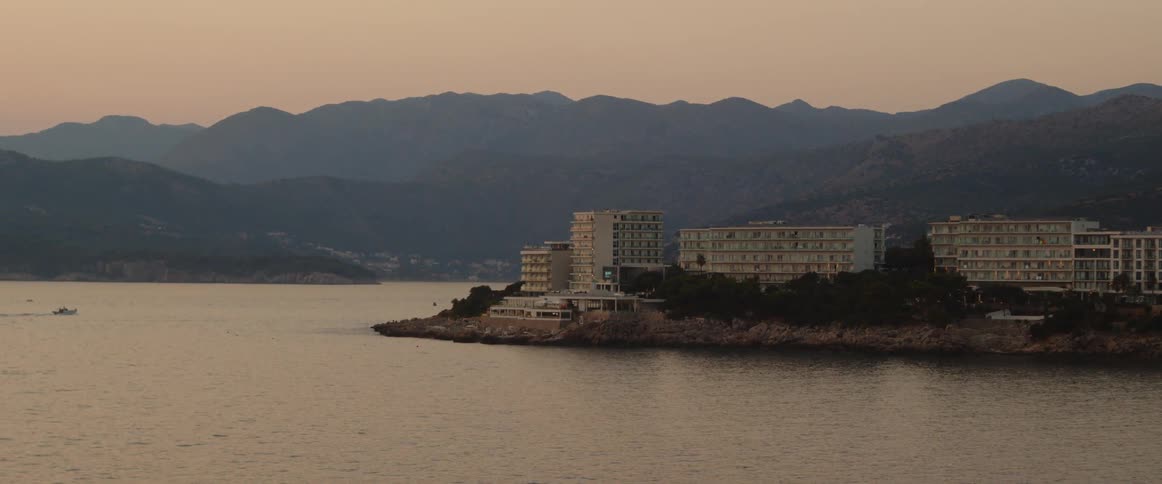 Boat moves toward apartment buildings and hotels on peninsula with fog covered mountain and town in background