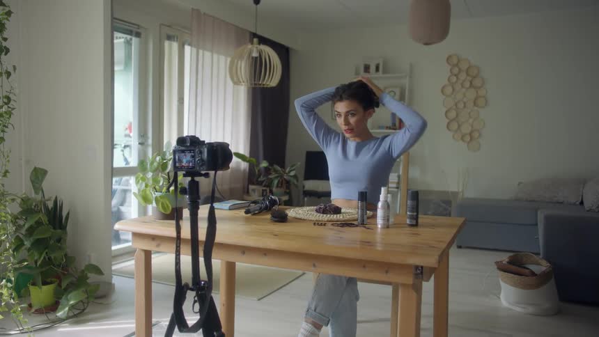 A woman in a room sitting on a chair at a table while talking to a camera with a wall and a door in the background