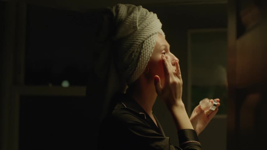A woman with a towel on her hair is putting cream on her face