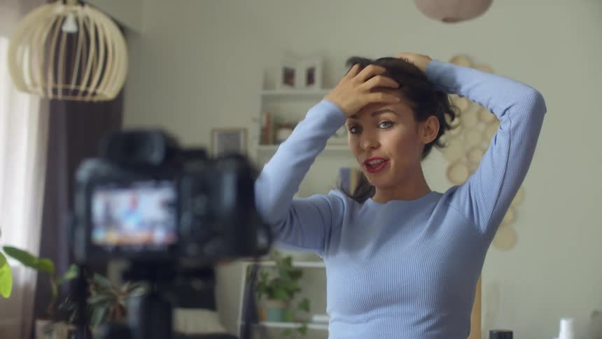 A woman films herself with a camera on a tripod while she demonstrates hair styles