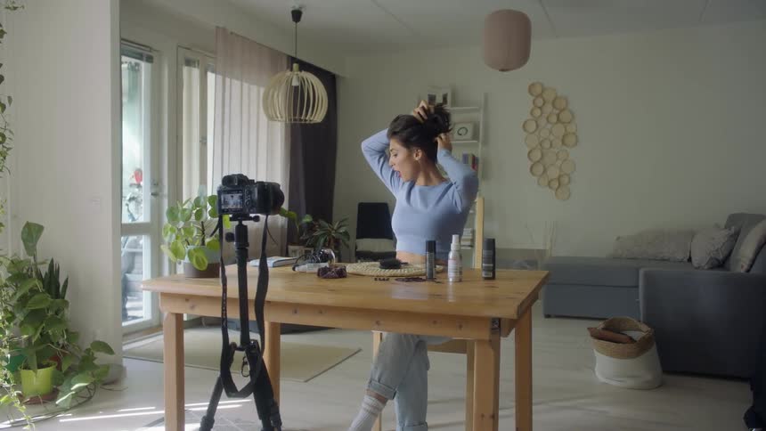 A woman is sitting at a table fixing her hair for the video camera in front of her while she is talking