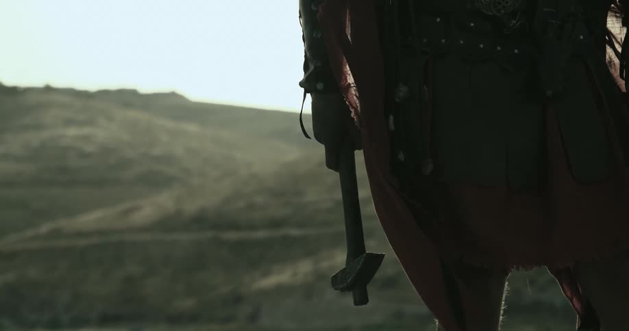 A man stands outside near tall hills holding a hammer and wearing medieval clothing