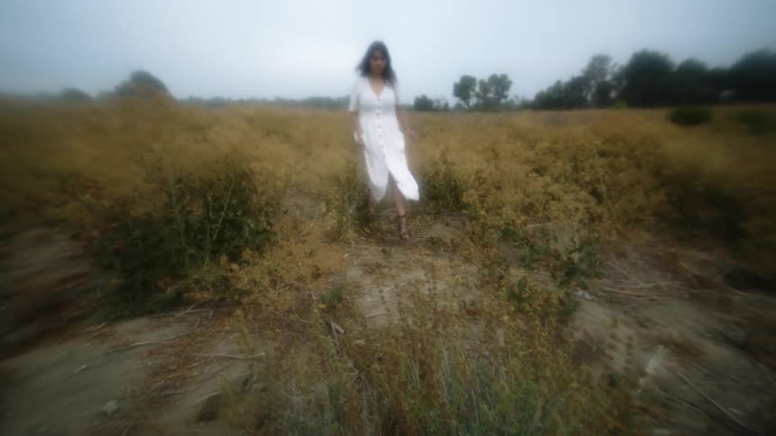 Woman wearing a white dress walks across field with woods in background
