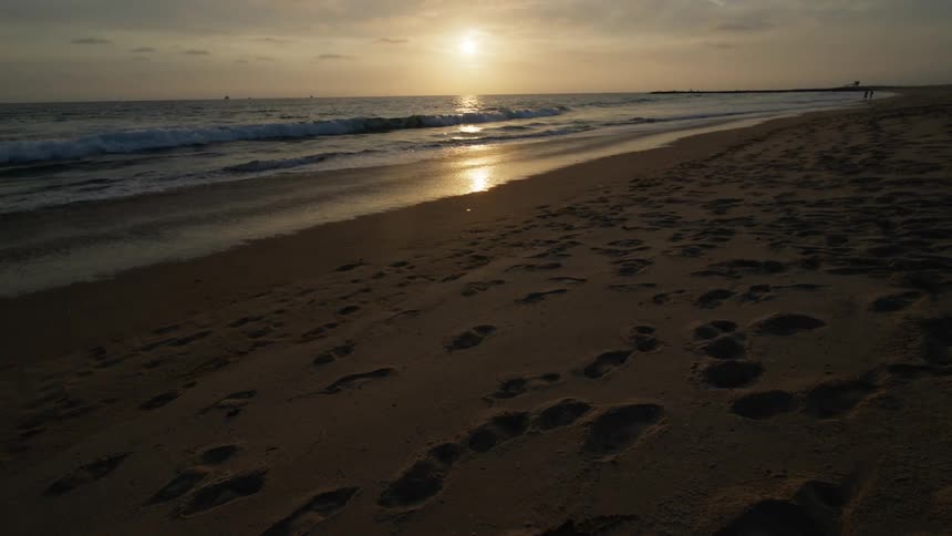 Myriad of footsteps cover sandy beach with people in the distance as water reflects sunshine