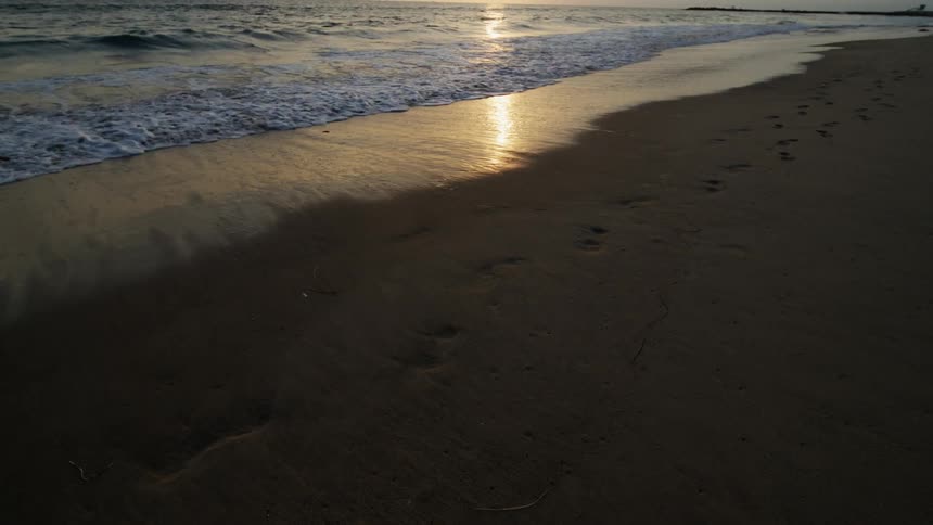 Waves roll over a beach as the reflection from the setting sun shines on the water