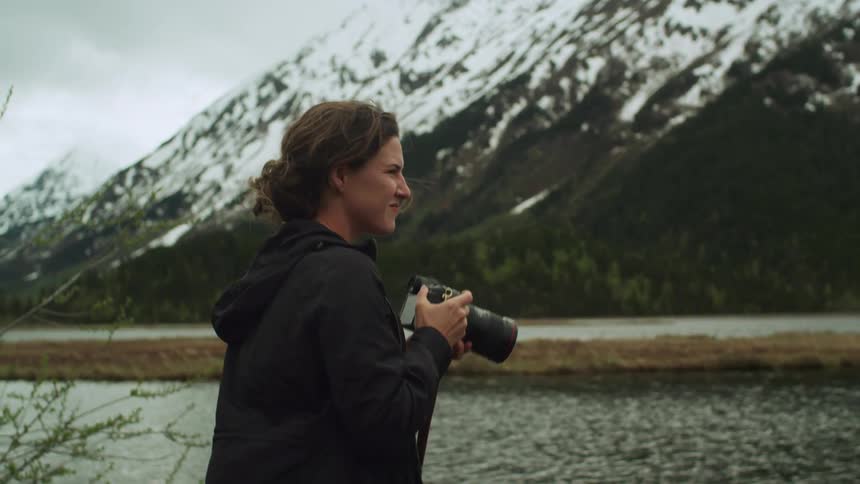 A female photographer stands next to a river near a snow covered mountain as she holds her camera to her eye