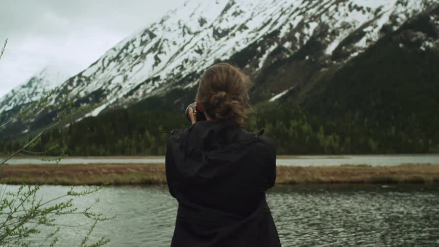 A female photographer aims her camera while standing outdoors next to a snow covered mountain and river