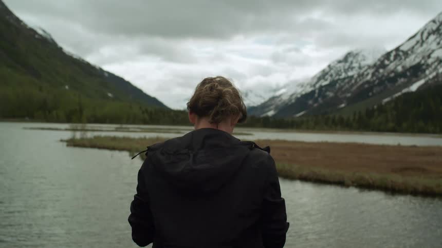 Female photographer with brown hair and black jacket takes photographs of lake with snowy high mountains against the cloudy sky