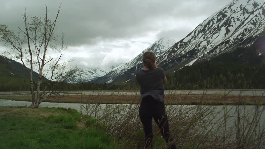 A woman takes a picture of snow clad mountains visible at a distance