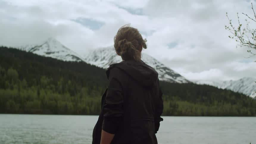 Woman stands on shore looking around with forested slope and snow covered mountains across water
