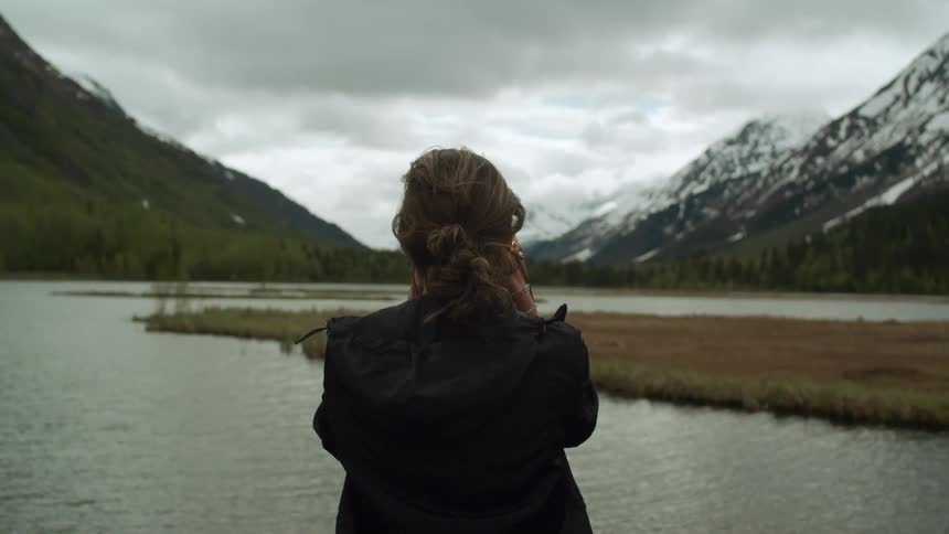 A woman stands still looking at a river that flows between mountains