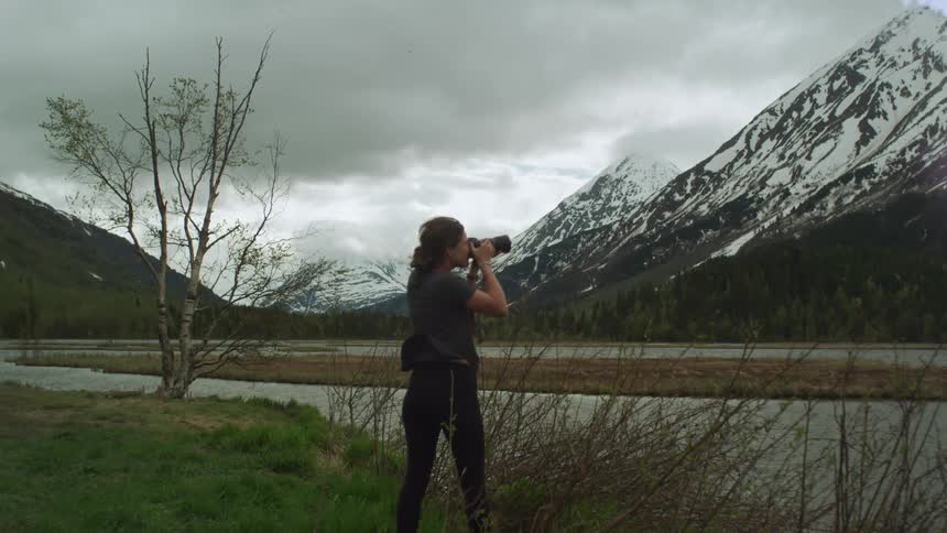 A woman with a camera taking pictures with a mountain range the sky and clouds in the background