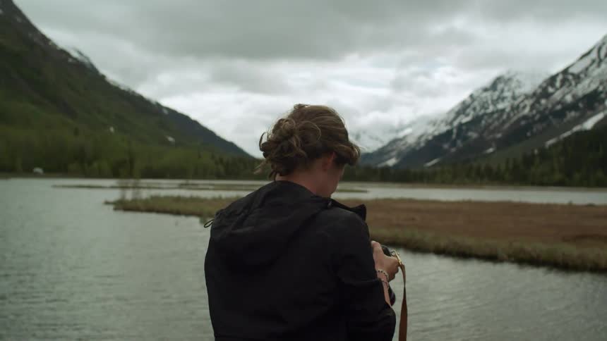 A photographer takes a photograph of a winding river in a mountain valley on a cloudy day before checking her photos on the cameras screen