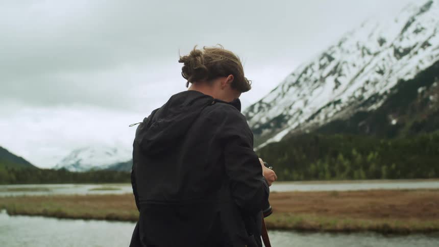 A female photographer is take a photo in a valley with snowy mountains on a breezy overcast day