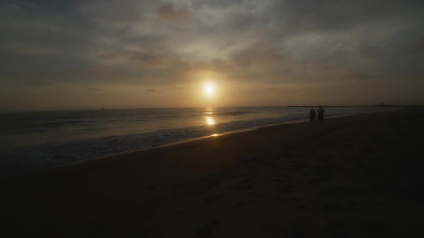Two people watch waves roll over a beach as the reflection from the setting sun shines on the water