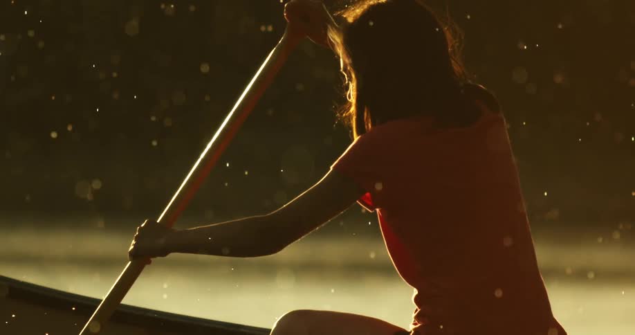 A woman uses a long paddle to row a canoe that she is in while it lightly rains