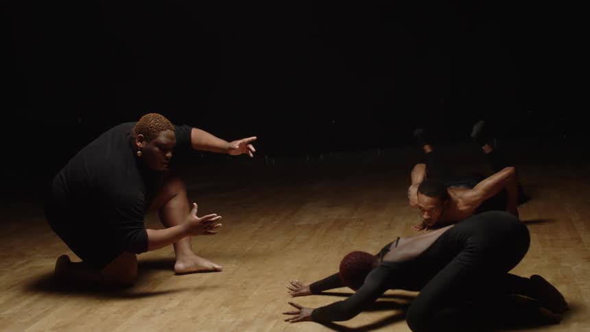 Three dancers perform modern dance routine in lit area of dark wood floored dance studio