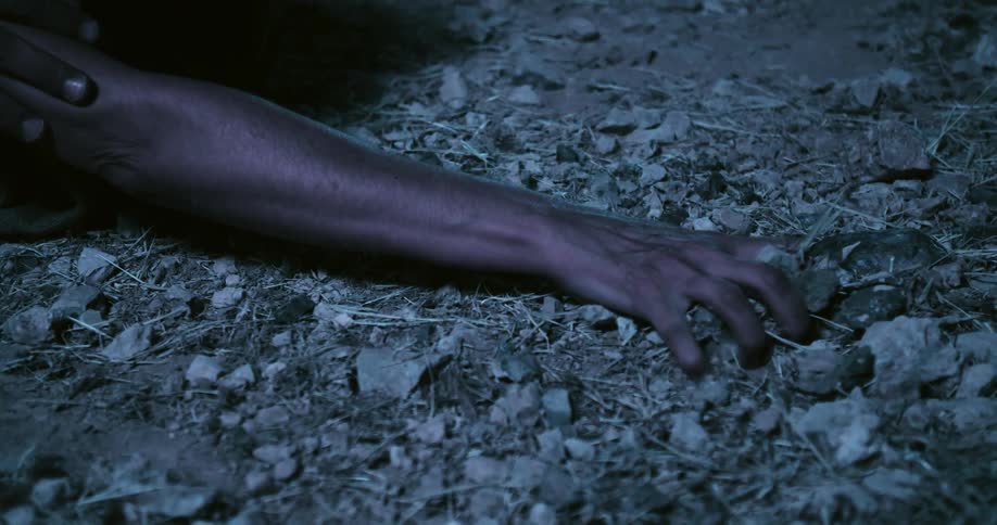 A person hesitantly reaches outward to grab a handful of rocks and dirt from the ground they are kneeling on