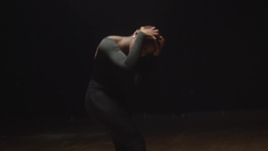 A woman in a black leotard twists around and grabs her face as she dances in a studio